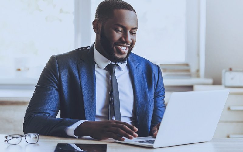 Young Beard Afro-american Man Working with Laptop Sitting at Table. Freelancer at Home. Work Concept. Web Surfing. Using Digital Device. Modern Workplace. Laptop on Desk. Businessman at Home.